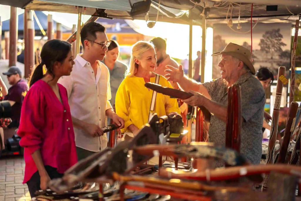 Local vendor explaining Aboriginal artifacts and boomerangs to tourists at an Australian cultural market stall.