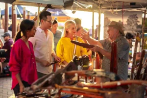 Local vendor explaining Aboriginal artifacts and boomerangs to tourists at an Australian cultural market stall.